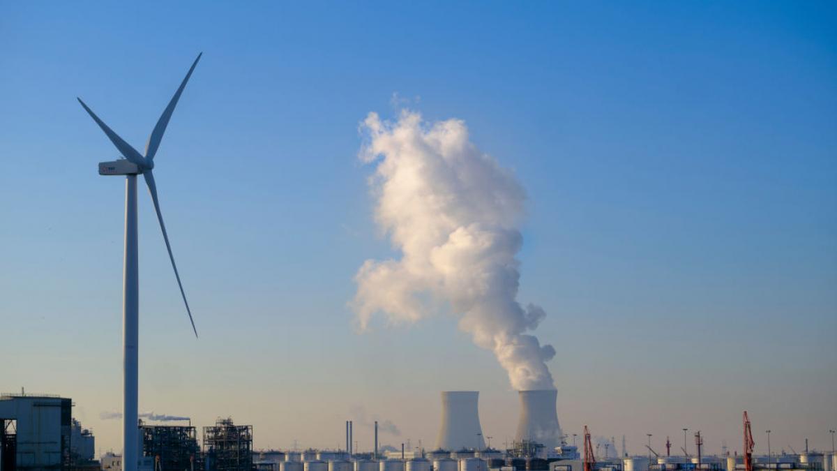 A picture of a windmill and a power plant, with white smoke coming out of the top of it. Blue skies