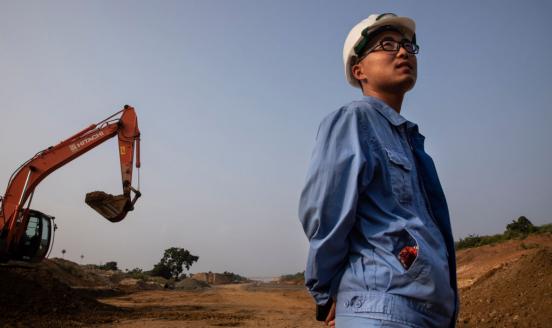 Photo of worker wearing glasses and blue overalls in front of a yellow excavator