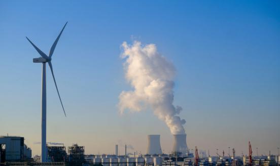 A picture of a windmill and a power plant, with white smoke coming out of the top of it. Blue skies