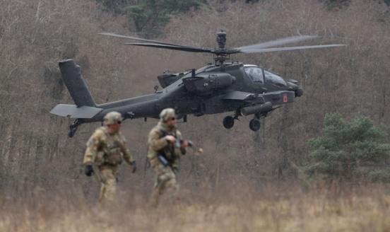 Two soldiers in camouflage with a helicopter taking off in the background.