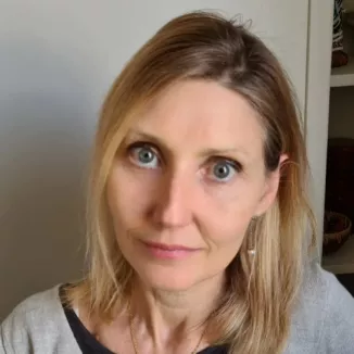 Headshot of a blonde woman with a blue eyes. She has a side parting and looks straight into the camera.