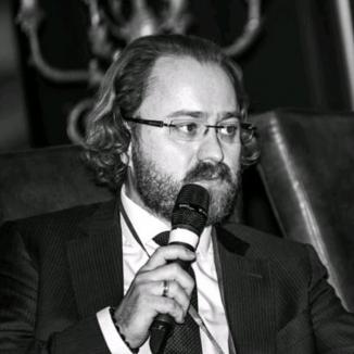 A man with beard and classes, holding a mic and talking