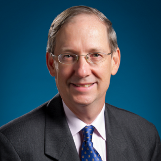 A man with glasses, in a grey suit, with a blue tie , smiling at the camera