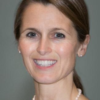 A woman with brown hair in a ponytail, in white blouse and pearl necklace smiling at the camera