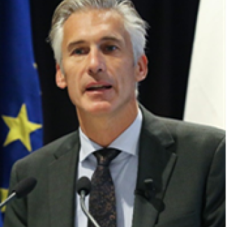 A man with grey hair, in dark green suit, speaking into the microphones,EU flag in the background