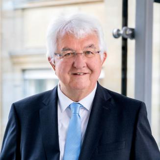 a man with grey hair, square glasses, in a black jacket, a white shirt and a bright blue tie