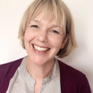 a woman with a short blonde hair wearing a burgundy cardigan and a grey shirt, smiling at the camera